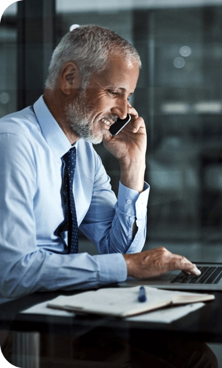 Middle aged white man with white hair wearing a dress shirt and tie, talking on a cell phone and focusing on a laptop screen.